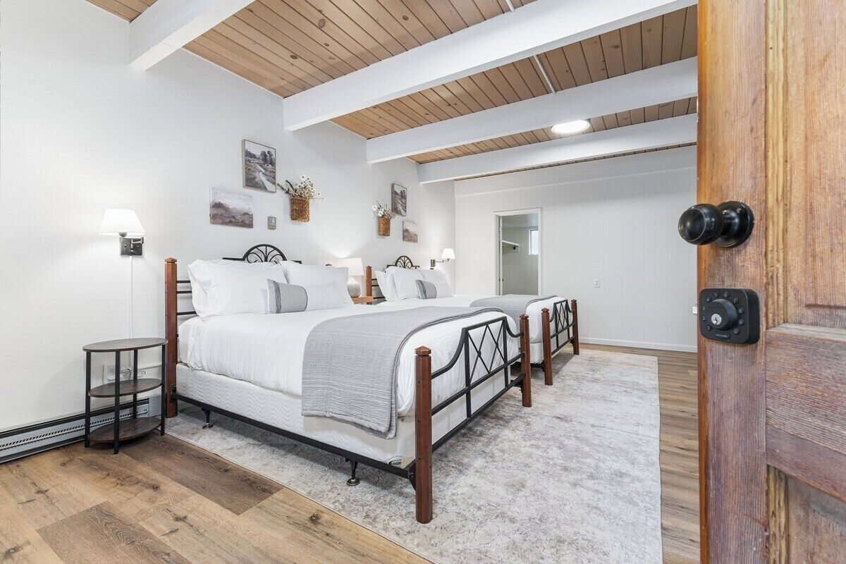 A bedroom with a wooden ceiling, two beds, bedside tables, wall-mounted lamps, and a door with a lock.