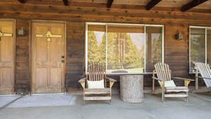 A log cabin with two wooden doors labeled "HI" and "ANG R", a large window with a view of trees, and two Adirondack chairs outside. - Camp Nelson Lodge | Room 4 | Sierra Nevada Retreat (Springville)