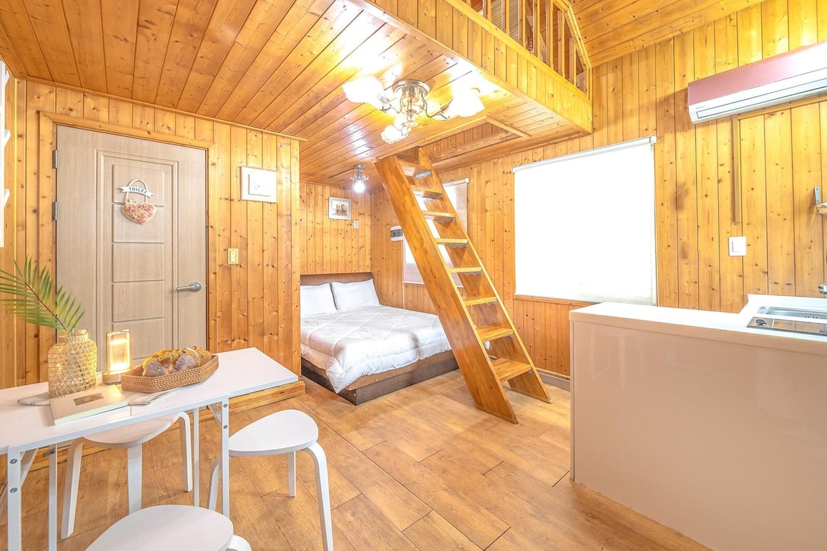 Ein Holzhütten-Innenraum mit Bett, Essbereich mit Stühlen, einer Küche inklusive Spüle und einer Leiter zu einer erhöhten Ebene.