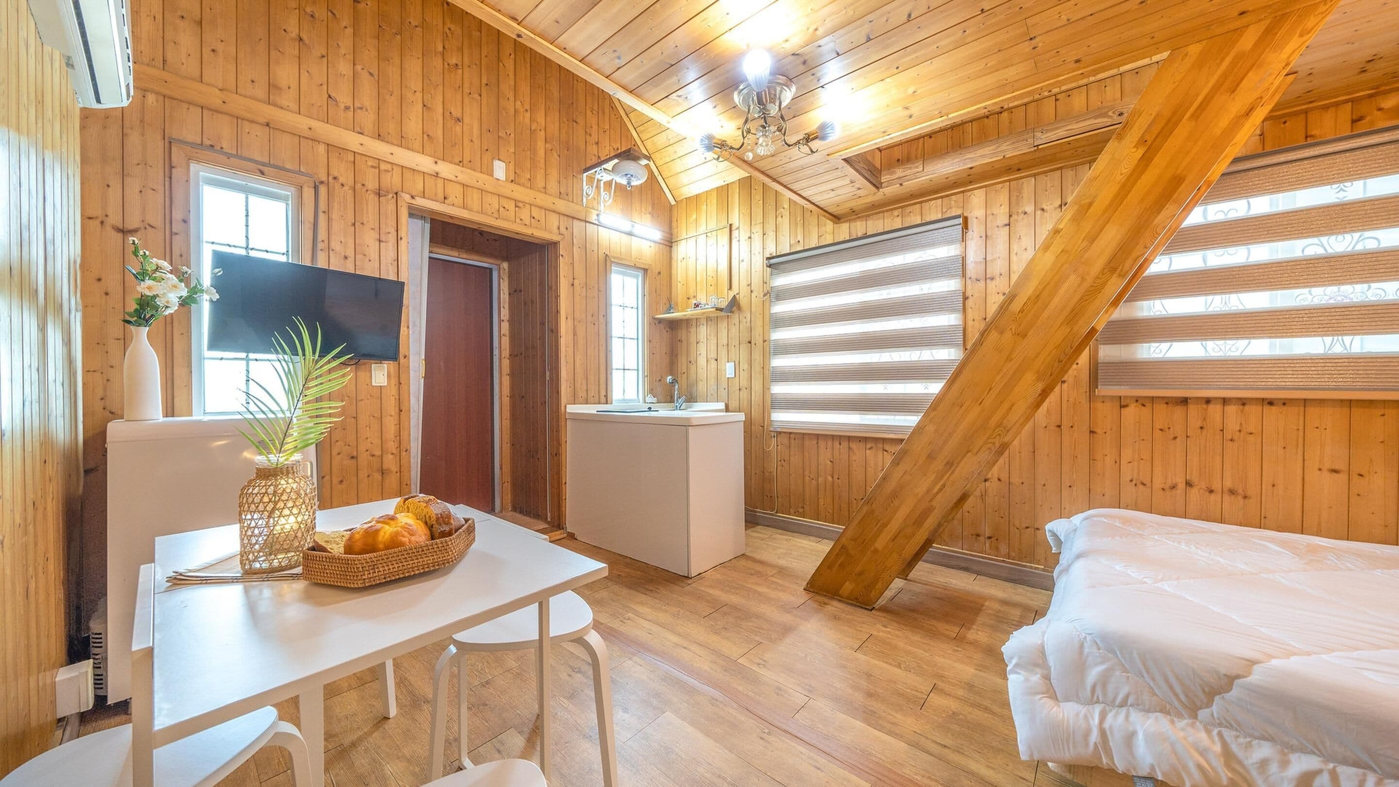 Puuinen mökkihuone, jossa on sänky, pieni keittokomero, pöytä, jossa on maljakko ja leipää, sekä seinällä oleva televisio.