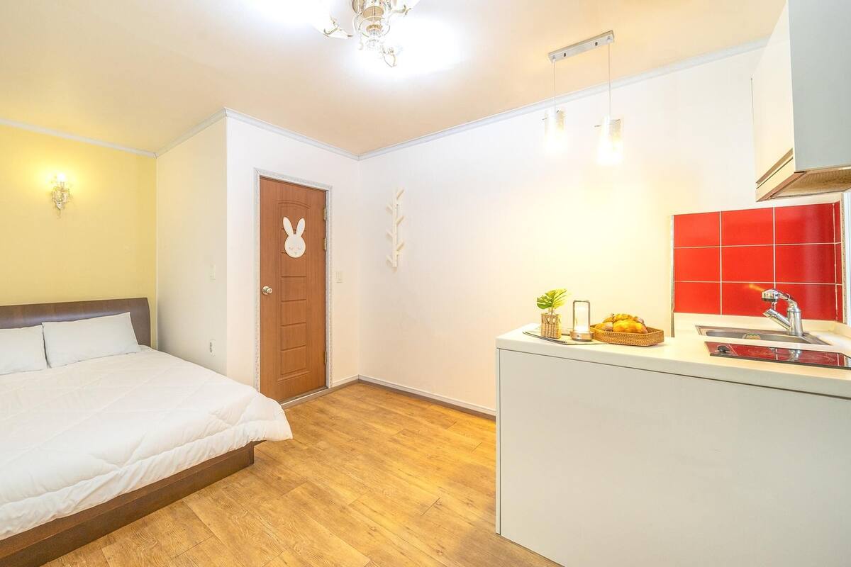 Ein Schlafzimmer mit einem Bett, einer Holztür mit Hase und einer kleinen Küchenzeile mit roter Spritzschutzwand sowie einem Holzboden.