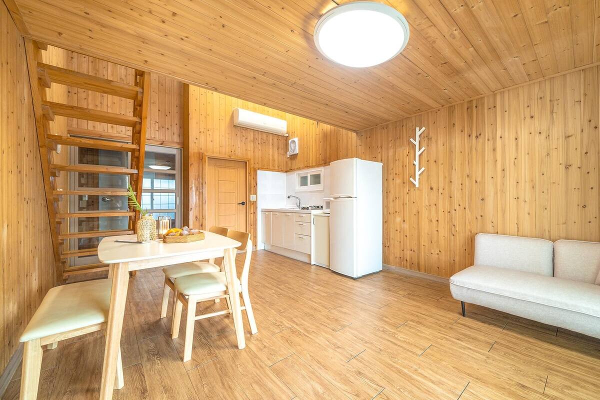Ein gemütlicher Raum mit Holzwänden, einem weißen Sofa, einem Esstisch mit Stühlen, einer Küchenzeile und einer Deckenleuchte.