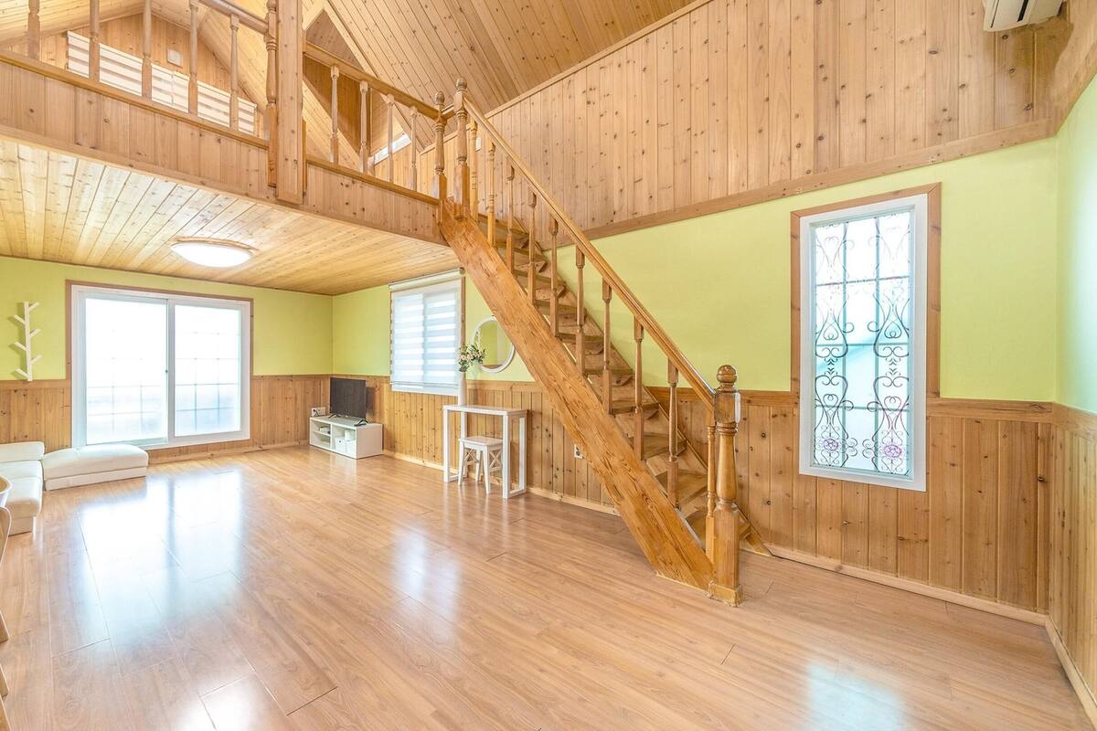 Ein geräumiges Zimmer mit Holzböden, einem großen Fenster, einer Treppe und einer Sitzecke mit Tisch und Stühlen.