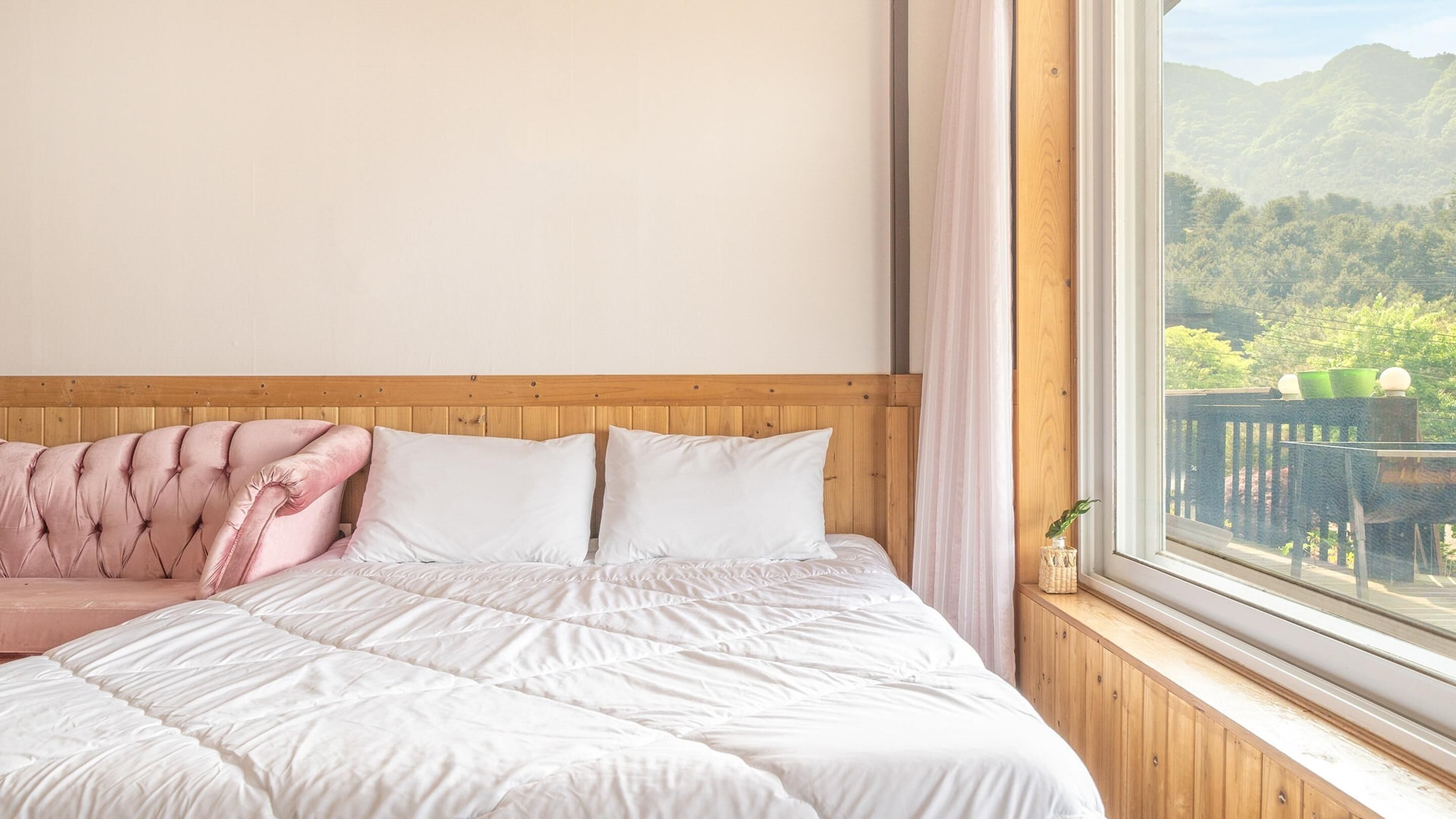 A bedroom with a large bed, a wooden headboard, a pink sofa, and a window with a view of greenery and mountains.