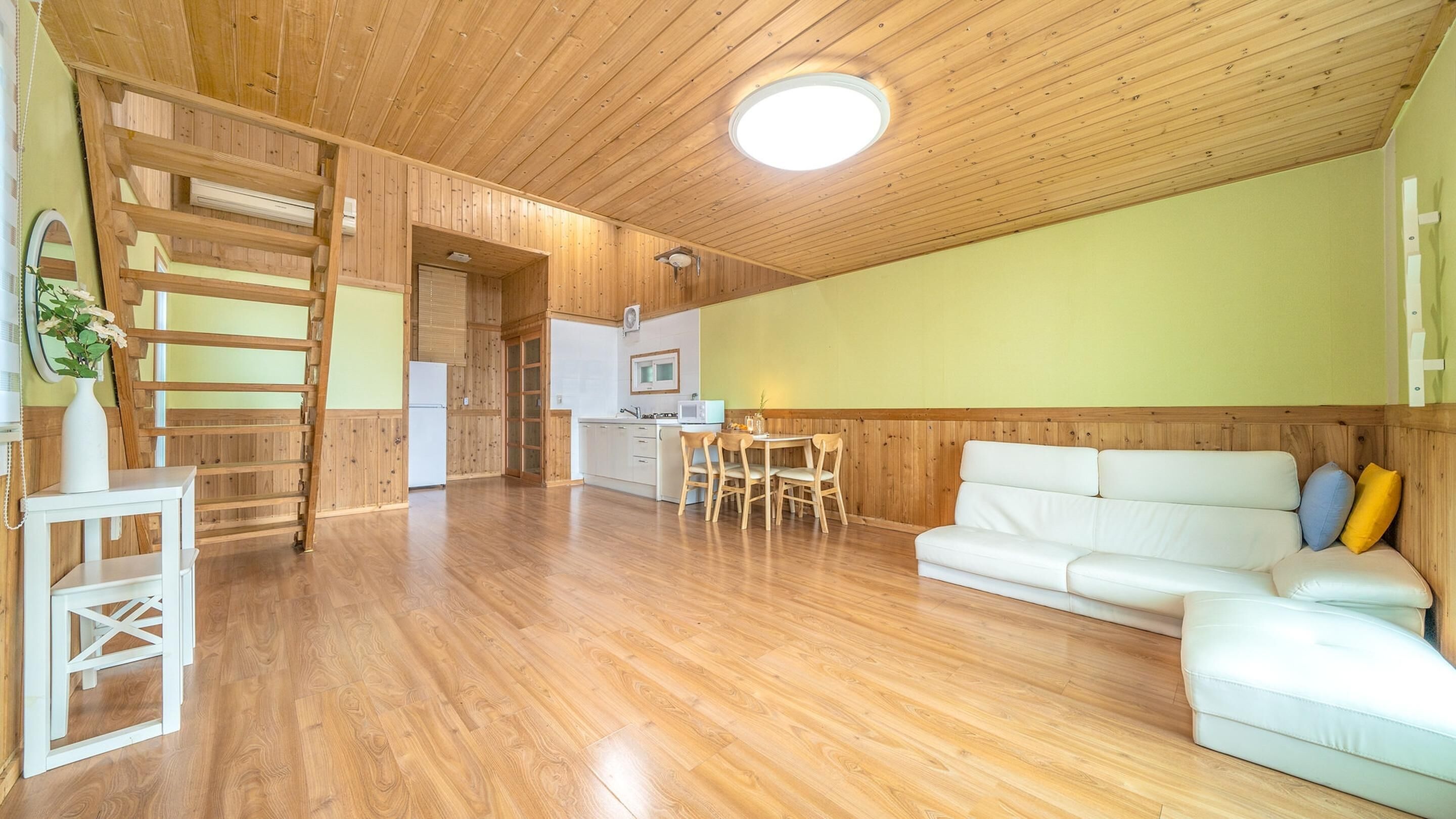 Amplia sala de estar con piso de madera, un sofá blanco, una mesa de comedor con sillas y una zona de cocina con microondas y refrigerador.