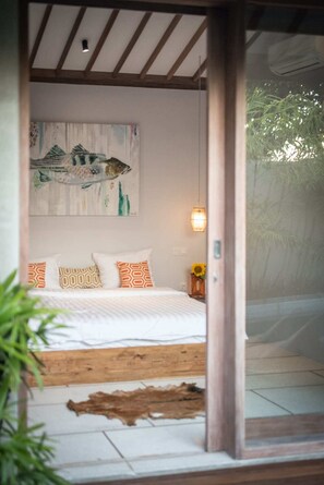 Ein Schlafzimmer mit Bett, Kissen, Wandbild, Pendelleuchte und Fenster mit Blick auf Grünflächen.