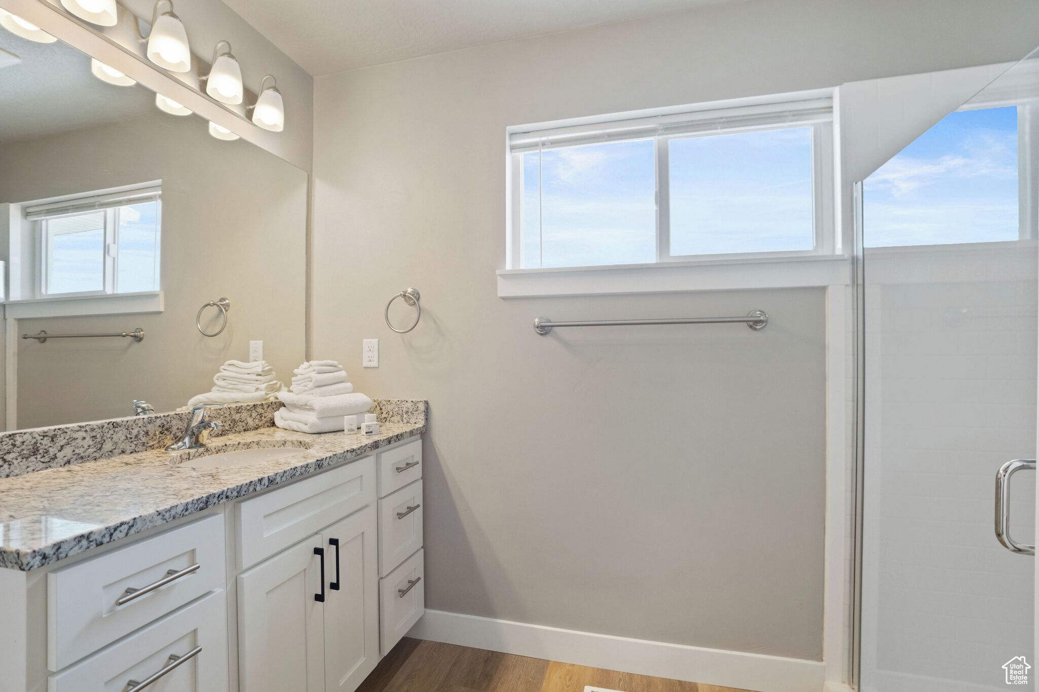 A bathroom with a granite countertop, white cabinets, a large mirror, and a shower with a window.