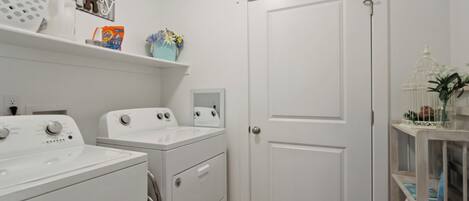 A laundry room with two white washing machines, a dryer, and a shelf with laundry supplies.