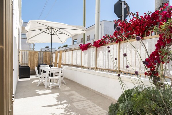 An outdoor patio with a table and chairs, a white umbrella, and a wall decorated with red flowers.