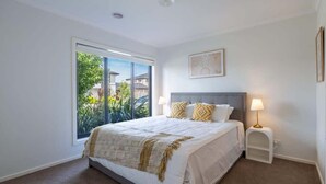 Un dormitorio con un ventanal grande, una cama con ropa de cama blanca, una mesita de noche con lámpara y un cuadro en la pared.