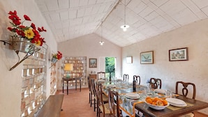 A dining area with a long wooden table set for six, surrounded by wooden chairs. A brick wall with a window and a hanging flower arrangement is visible. - Villa Ortensie Garden with Amazing View (Marliana)