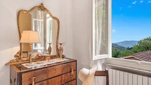 A wooden dresser with a mirror, a lamp, and a hat on a chair by a window with a view of greenery and mountains. - Villa Ortensie Garden with Amazing View (Marliana)