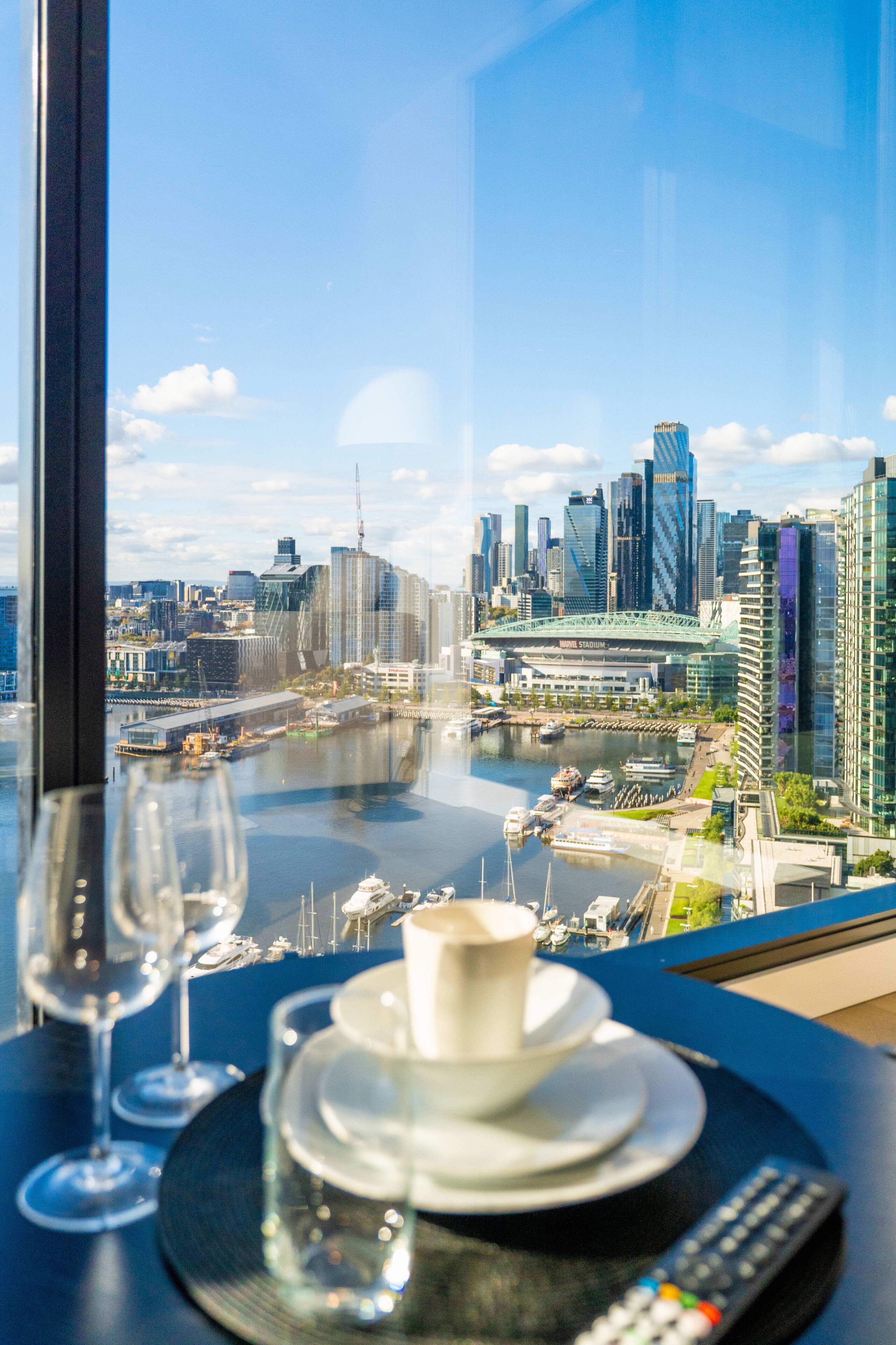 A table with a remote control, a cup, and wine glasses, overlooking a cityscape with modern buildings and a river.