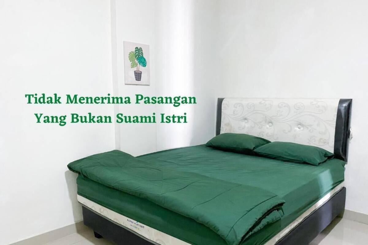 A neatly made bed with a green bedspread and matching pillows, a white headboard, and a small framed picture on the wall.
