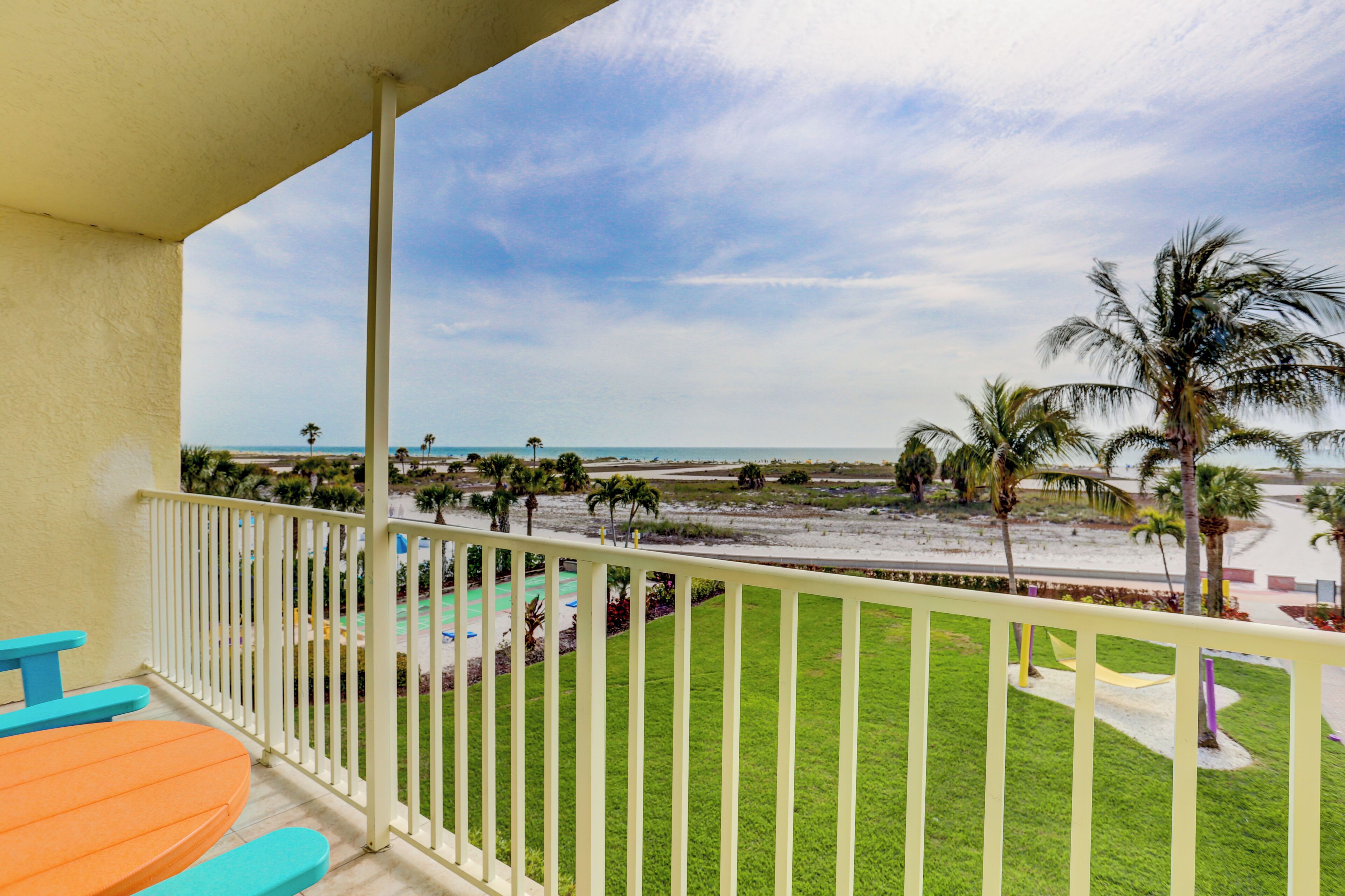 A balcony with a view of the beach, palm trees, and a playground.