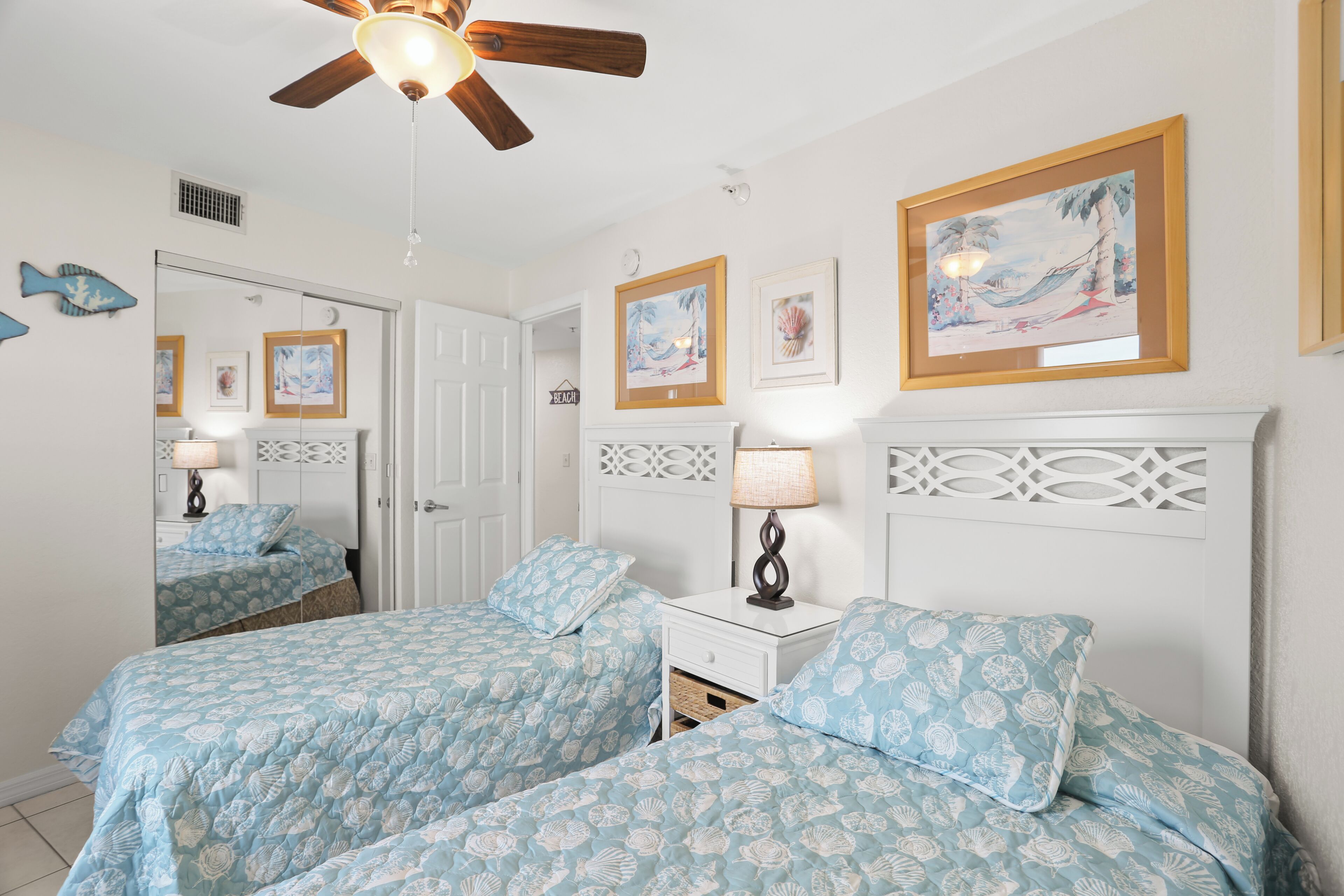 A bedroom with two beds, a ceiling fan, wall art, and a white headboard.
