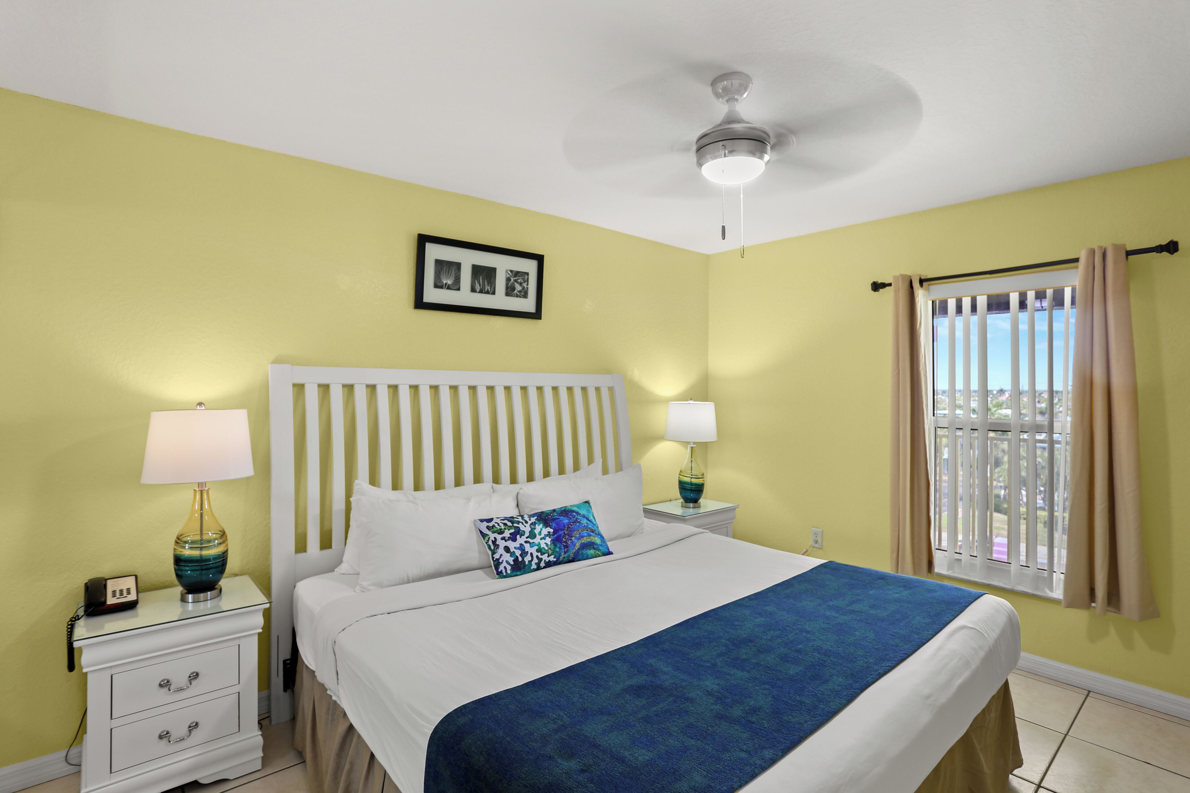 A bedroom with a bed, nightstands, a ceiling fan, a window with curtains, and a wall-mounted picture.