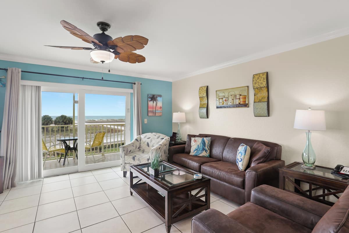 A living room with a ceiling fan, a glass coffee table, a brown leather sofa, and a view of the outdoors through a sliding glass door.