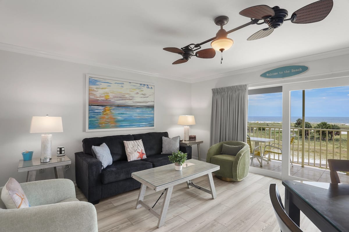 A living room with a sofa, coffee table, and chairs, a painting, a ceiling fan, and a balcony with a view.