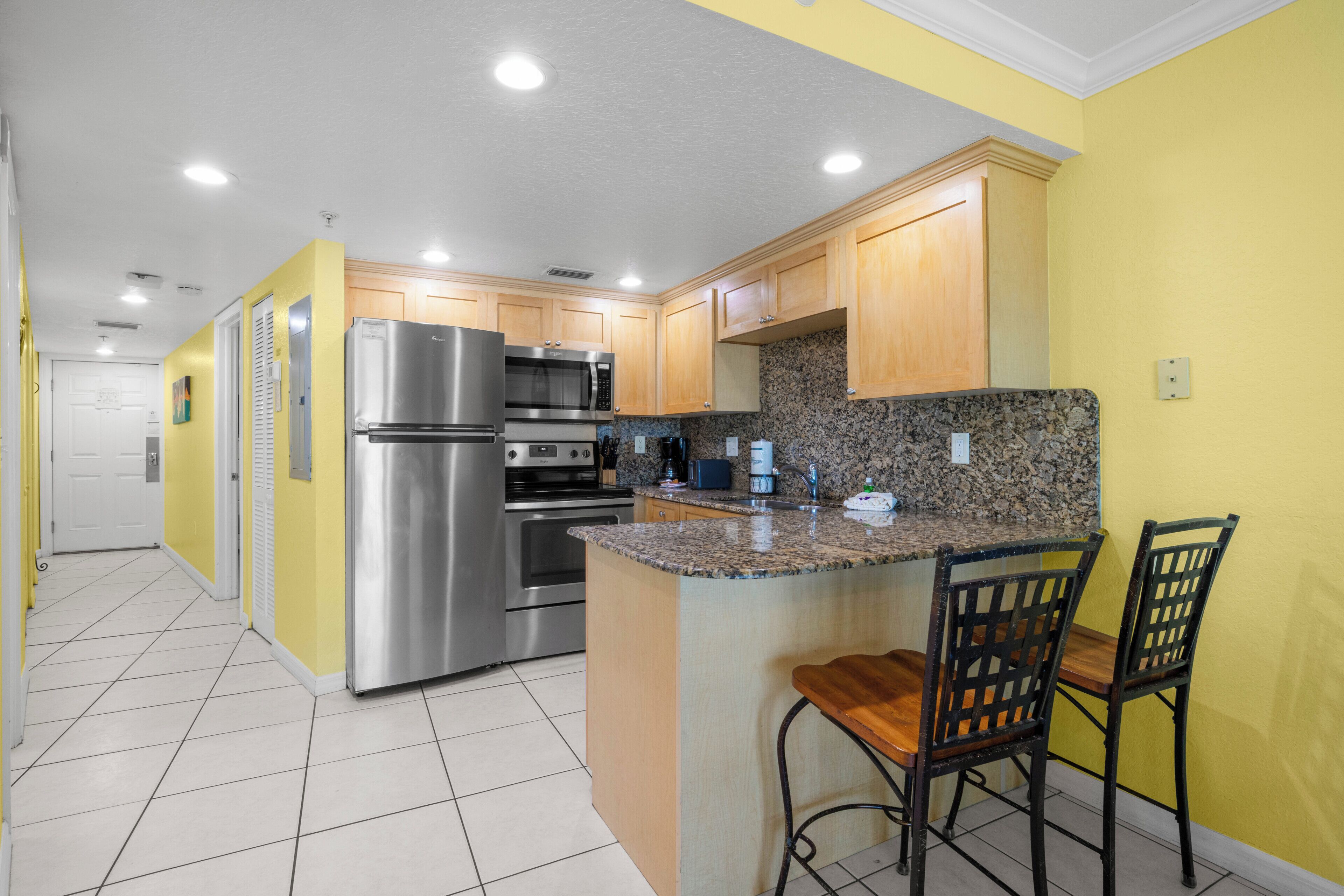 A kitchen with stainless steel appliances, granite countertops, wooden cabinets, and a bar area with two chairs.