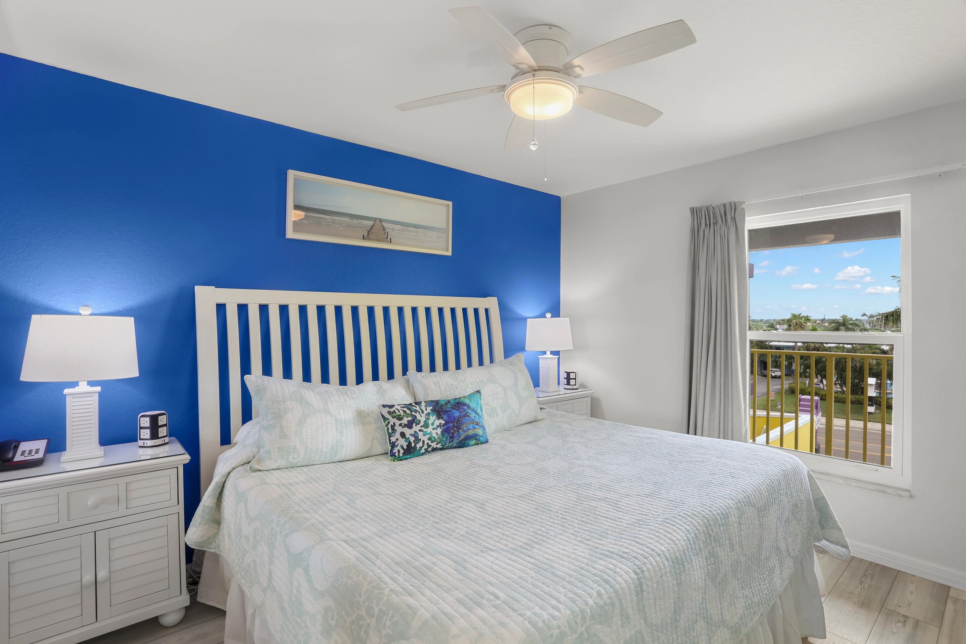 A bedroom with a bed, two bedside tables, a ceiling fan, a window with a view, and a painting on the wall.
