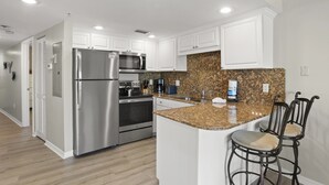 A modern kitchen with stainless steel appliances, white cabinetry, granite countertops, and a bar area with two stools. - South Beach Condo Hotel (Treasure Island)