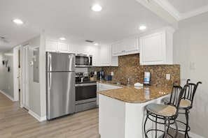 A modern kitchen with stainless steel appliances, white cabinetry, granite countertops, and a bar area with two stools.