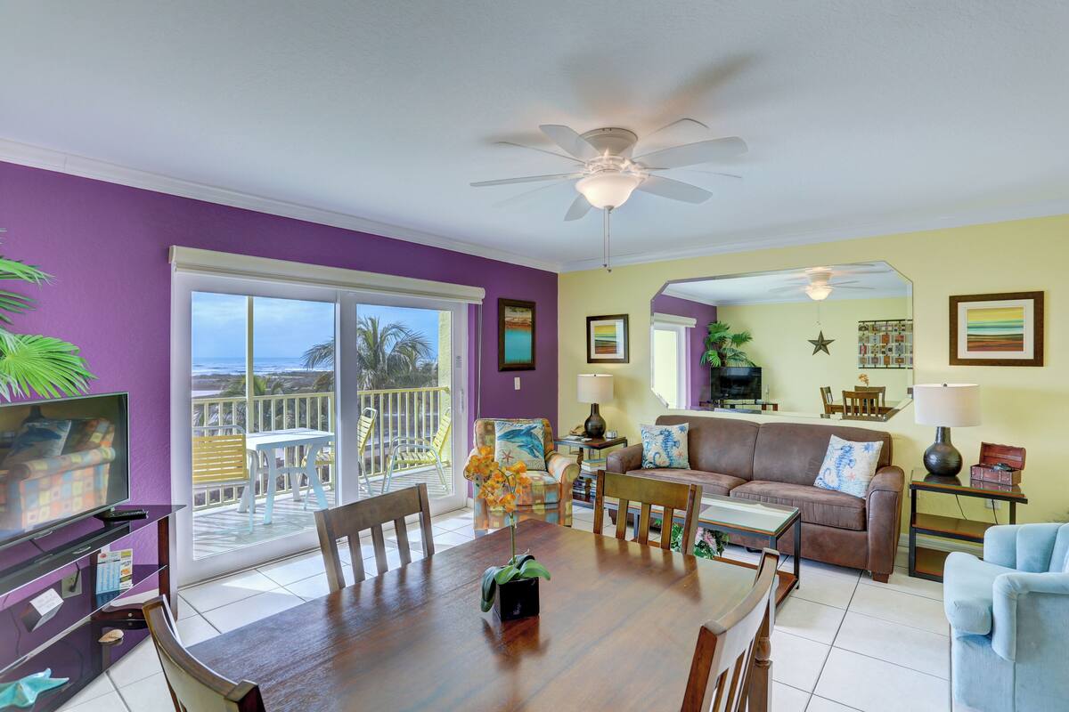 A living room with a dining table, chairs, a sofa, and a TV. There is a balcony with a view of the ocean.