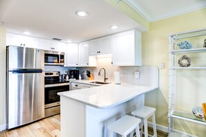 A modern kitchen with white cabinets, stainless steel appliances, a central island with seating, and a decorative shelf. - South Beach Condo Hotel (Treasure Island)