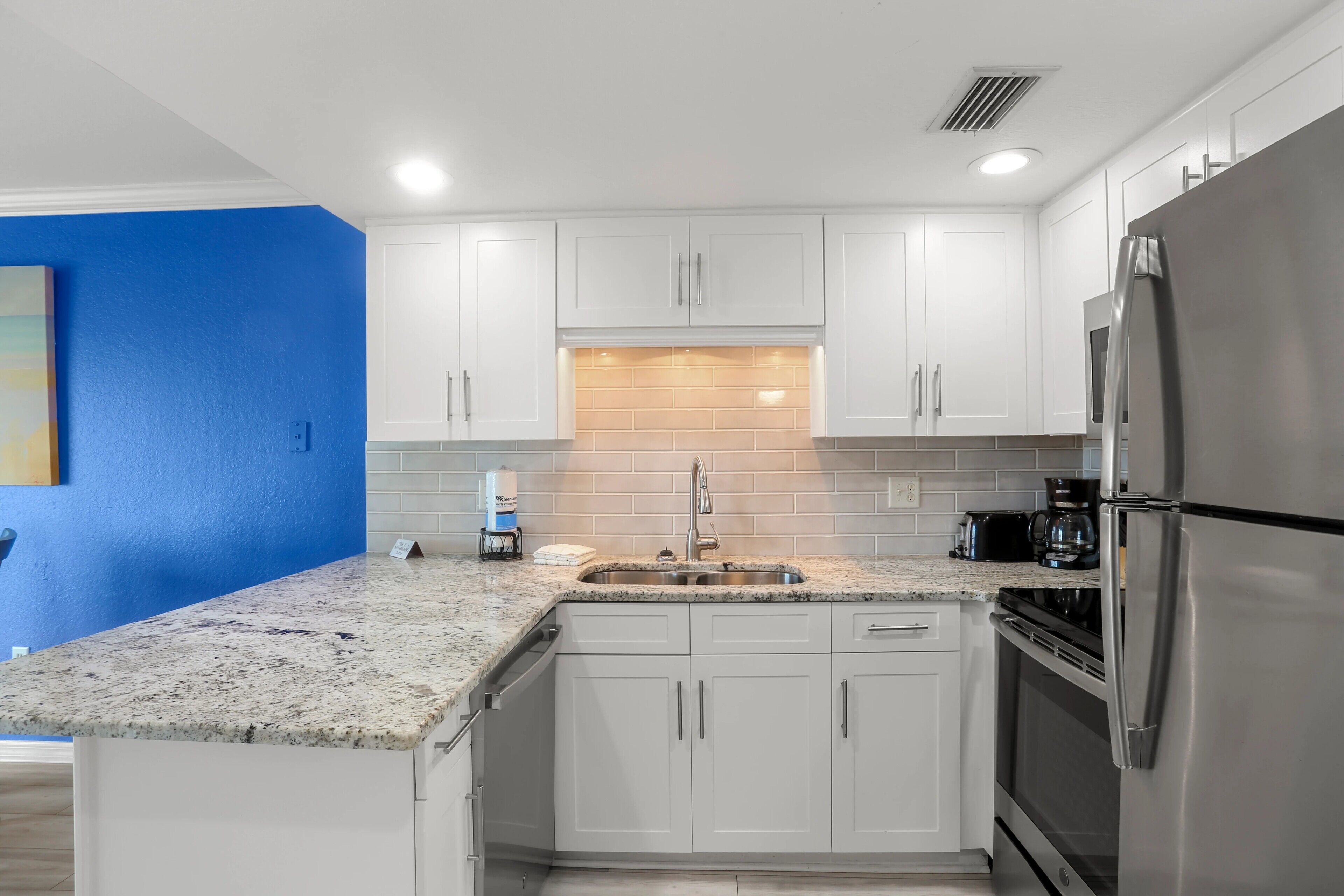Una cocina con armarios blancos, encimera de granito, refrigerador de acero inoxidable y cafetera.