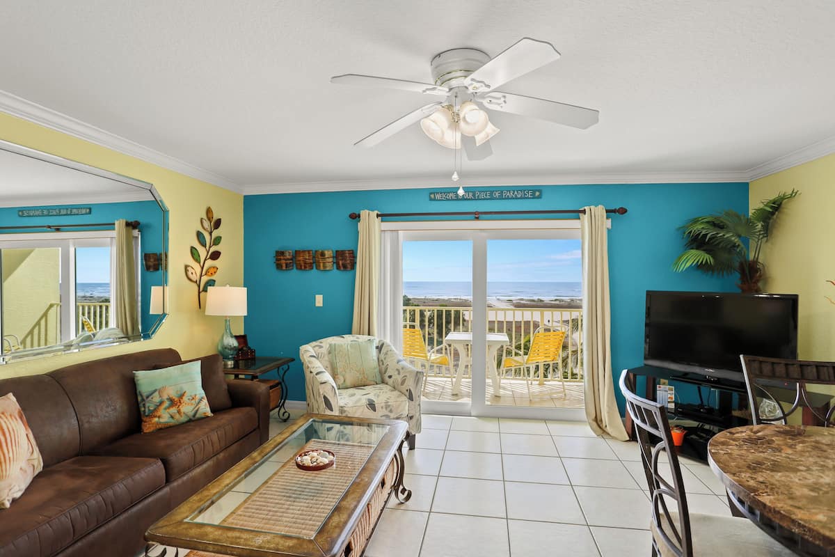 A living room with a sofa, coffee table, TV, and a balcony with yellow chairs.