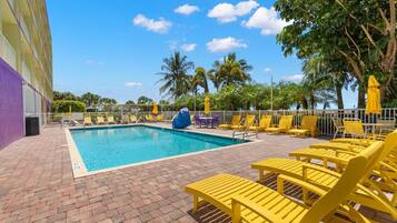 A swimming pool with yellow lounge chairs and umbrellas, surrounded by palm trees and a clear sky.