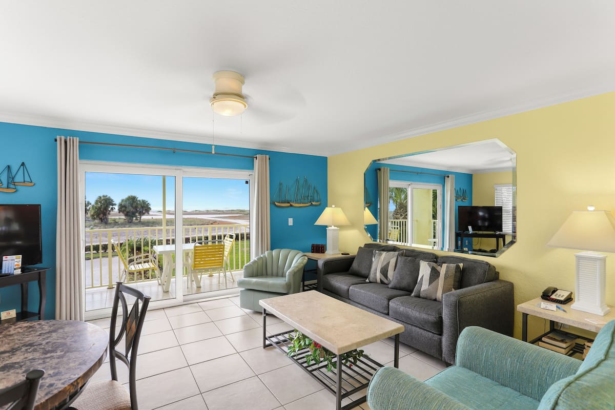A living room with a sofa, armchairs, a coffee table, a TV, and a balcony with outdoor furniture.