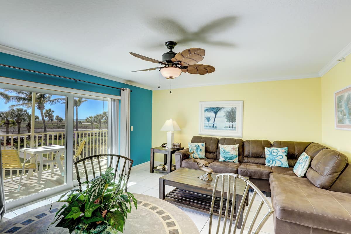 A living room with a ceiling fan, a sofa set, a coffee table, and a view of palm trees through large sliding glass doors.