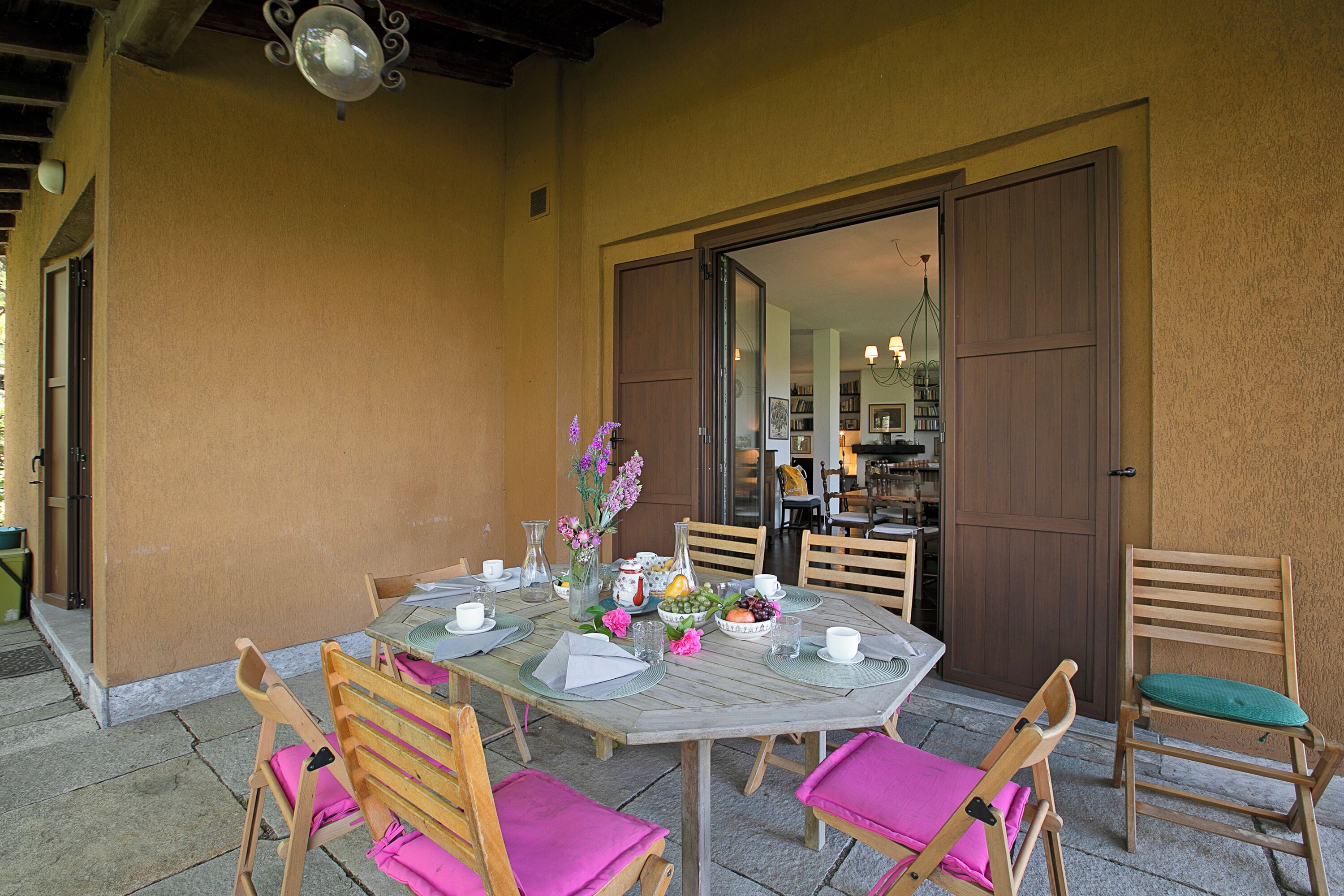 Un área de comedor al aire libre con una mesa dispuesta para cuatro personas, rodeada de sillas de madera con cojines rosados, un jarrón con flores y un cuenco de fruta.