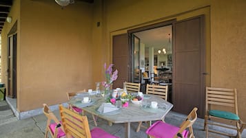 Un área de comedor al aire libre con una mesa dispuesta para cuatro personas, rodeada de sillas de madera con cojines rosados, un jarrón con flores y un cuenco de fruta.