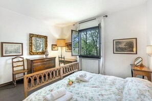 Un dormitorio con una cama de madera, un tocador, una silla y un ventanal con vistas a árboles.