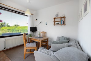 Une pièce comprenant une table à manger en bois, un canapé gris, un téléviseur, une plante et une fenêtre avec des stores.