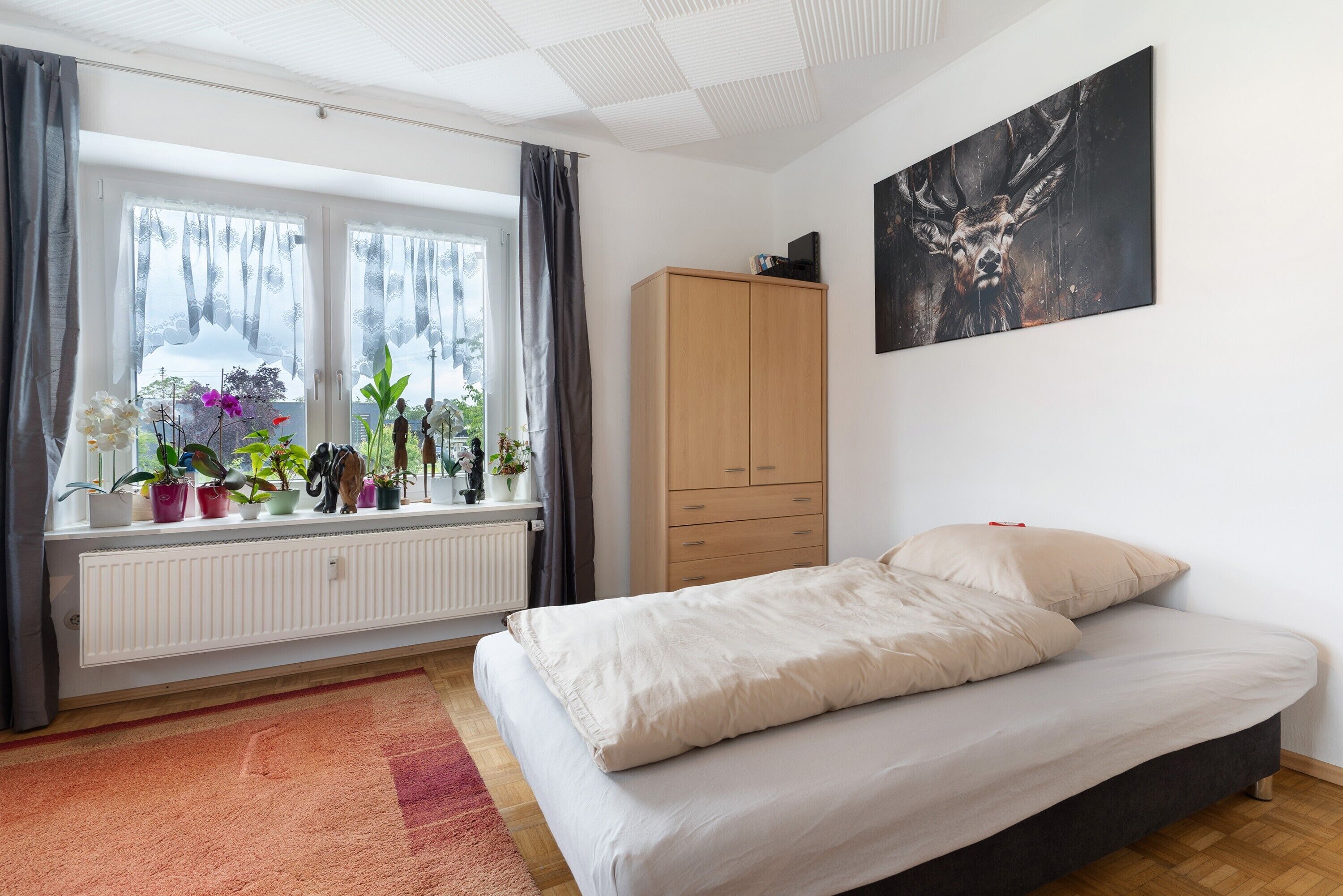A bedroom with a bed, a radiator, potted plants, a wardrobe, and a painting on the wall.