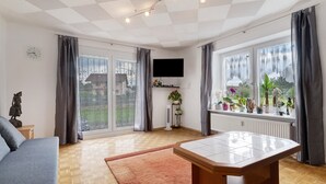 Ein modernes Wohnzimmer mit einer Couch, einem Esstisch, einem Fernseher und großen Fenstern mit Vorhängen.