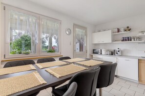 A dining area with a table set for six, a kitchen with white cabinets and appliances, and a view of greenery outside. - Apartment 'Ferienwohnung Am Brunnen' with Private Terrace and Wi-Fi (Durbach)