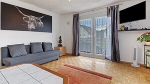 Ein Wohnzimmer mit einer Couch, einem Flachbildfernseher, einem großen Fenster mit Vorhängen und einer Decke mit geometrischem Muster.