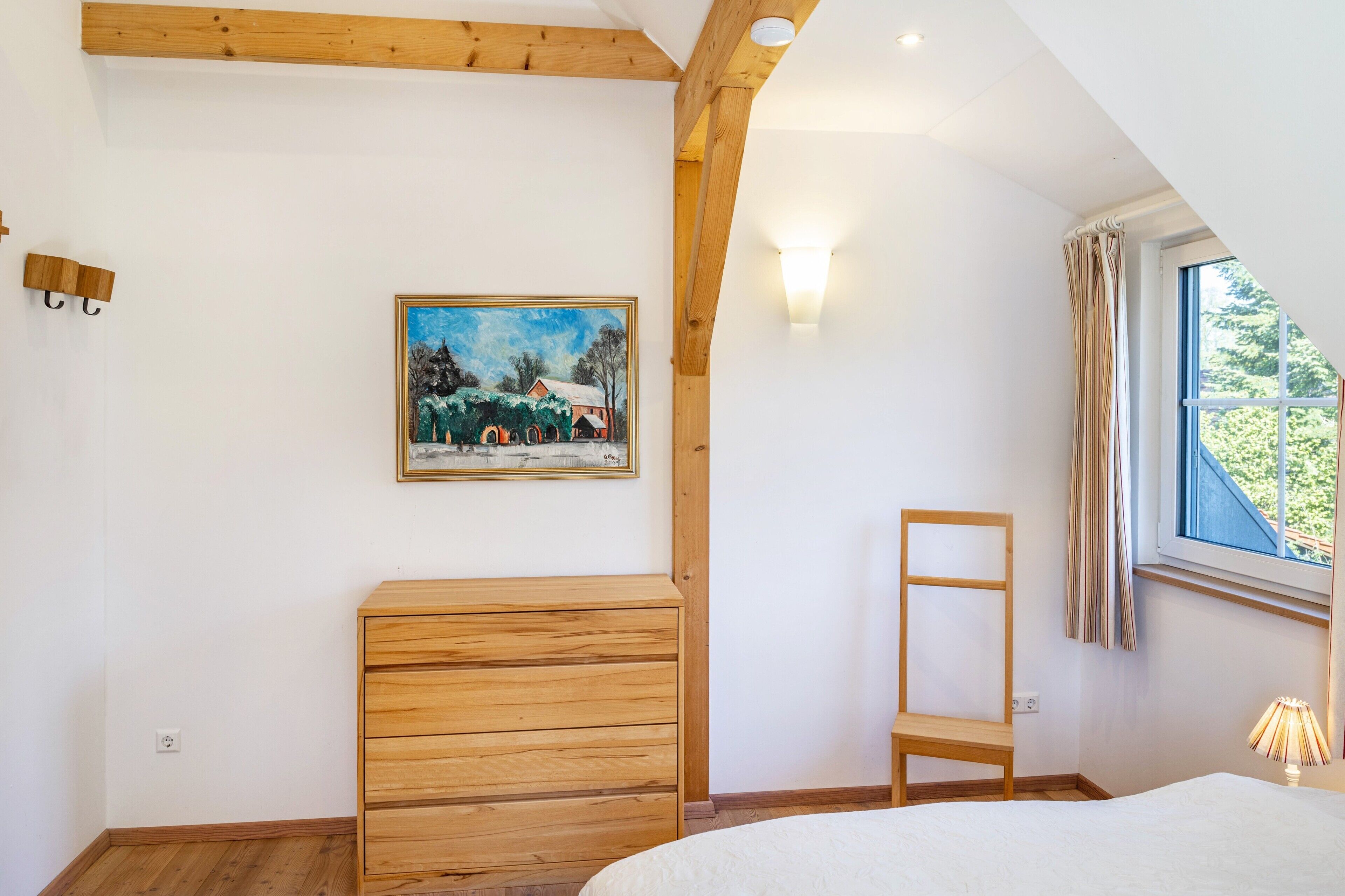 Une chambre à coucher avec une commode en bois, une chaise, un tableau, une fenêtre avec des rideaux et une lampe.