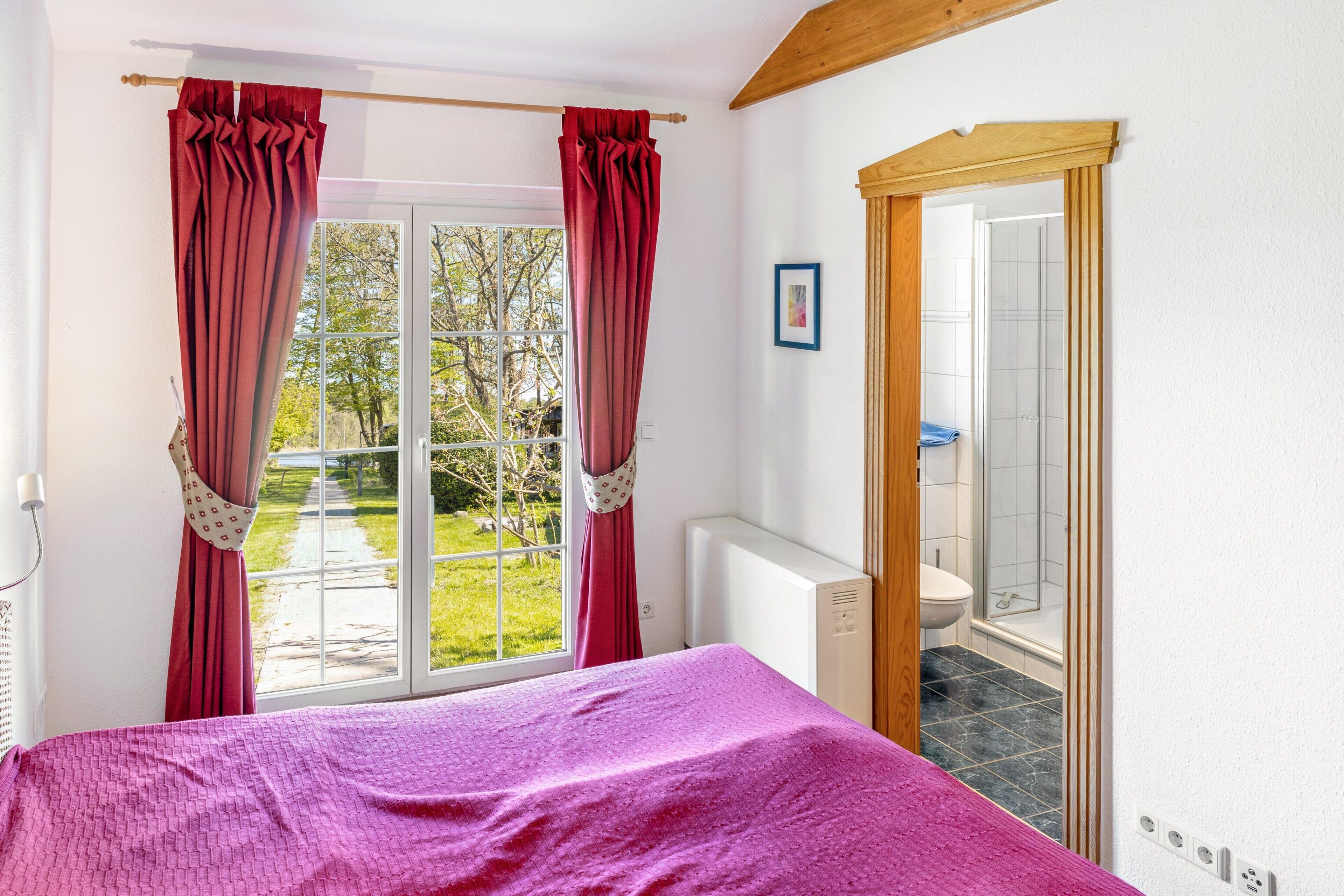 Un dormitorio con cama, ventana con cortinas, radiador y vista a un jardín y baño.