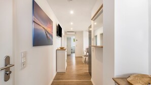 Un pasillo con piso de madera, una puerta con picaporte, un interruptor de luz en la pared, un televisor y vista a una habitación con una silla y una mesa.