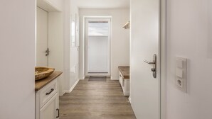 Un estrecho pasillo con piso de madera, una puerta blanca con picaporte, un pequeño banco y un cuenco de madera sobre una encimera.