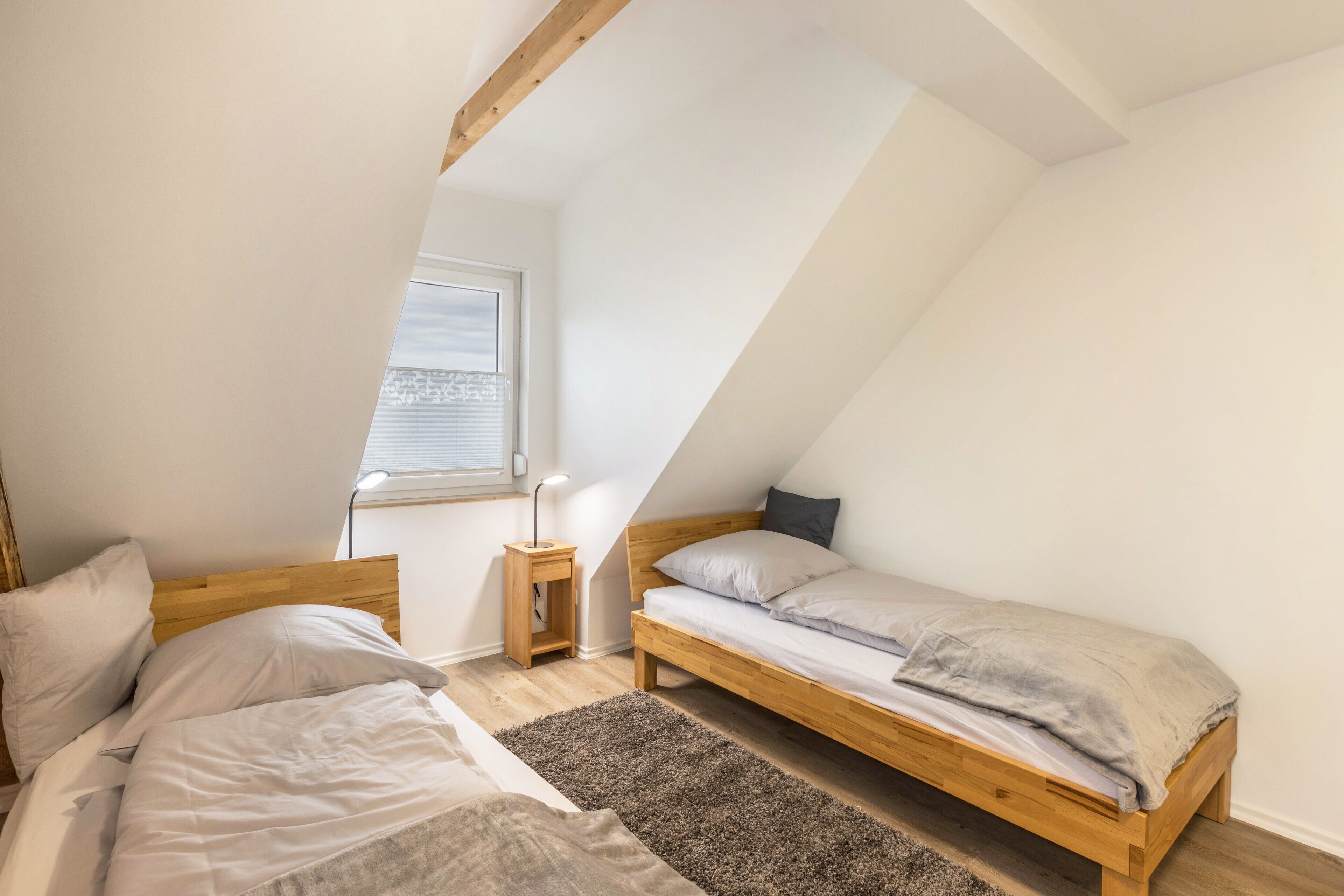 Un dormitorio con techo inclinado, una cama de madera, una mesita de noche, una lámpara y una ventana con persianas.
