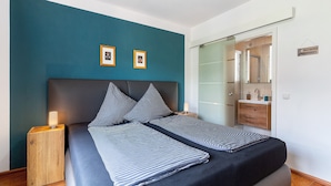 A bedroom with a bed, bedside tables, a nightstand, a mirror, and a bathroom visible through an open door.