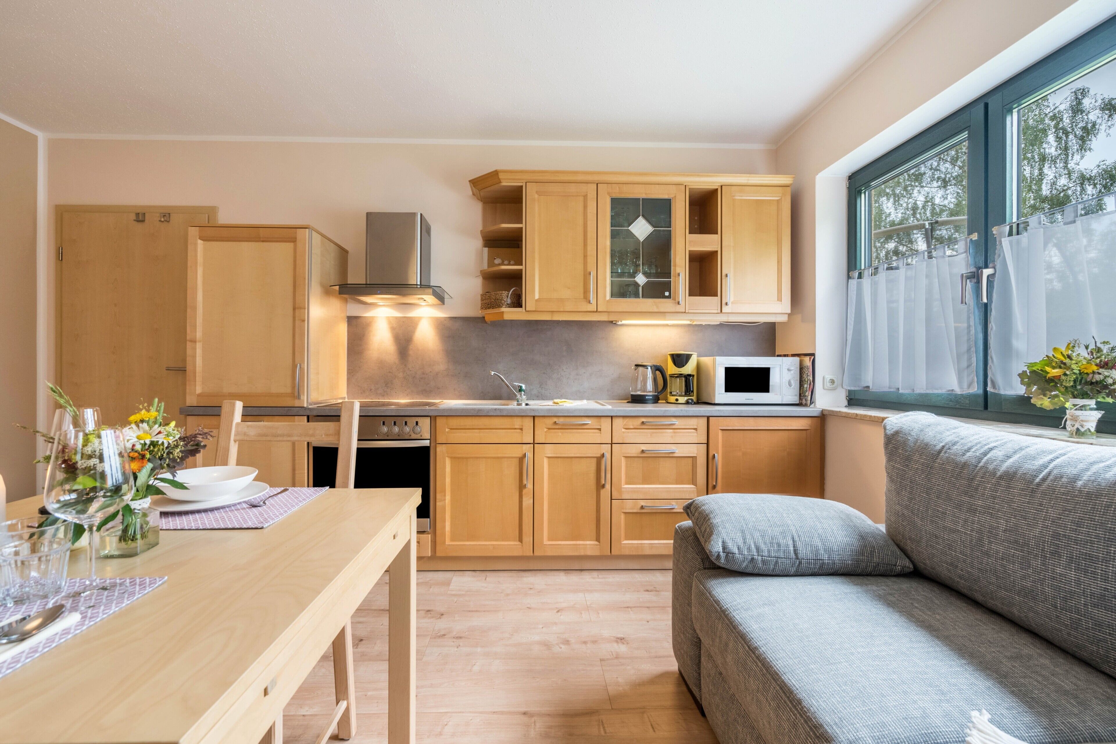 A compact kitchen with wooden cabinets, a dining table with a vase of flowers, and a sofa by a window.