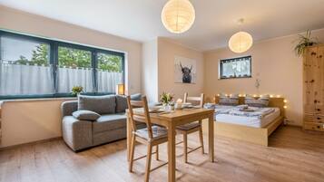 Ein modernes Zimmer mit einer Couch, einem Esstisch und einem Bett. Es gibt Fenster mit Vorhängen, ein an der Wand befestigtes Bild und eine Holzkommode.